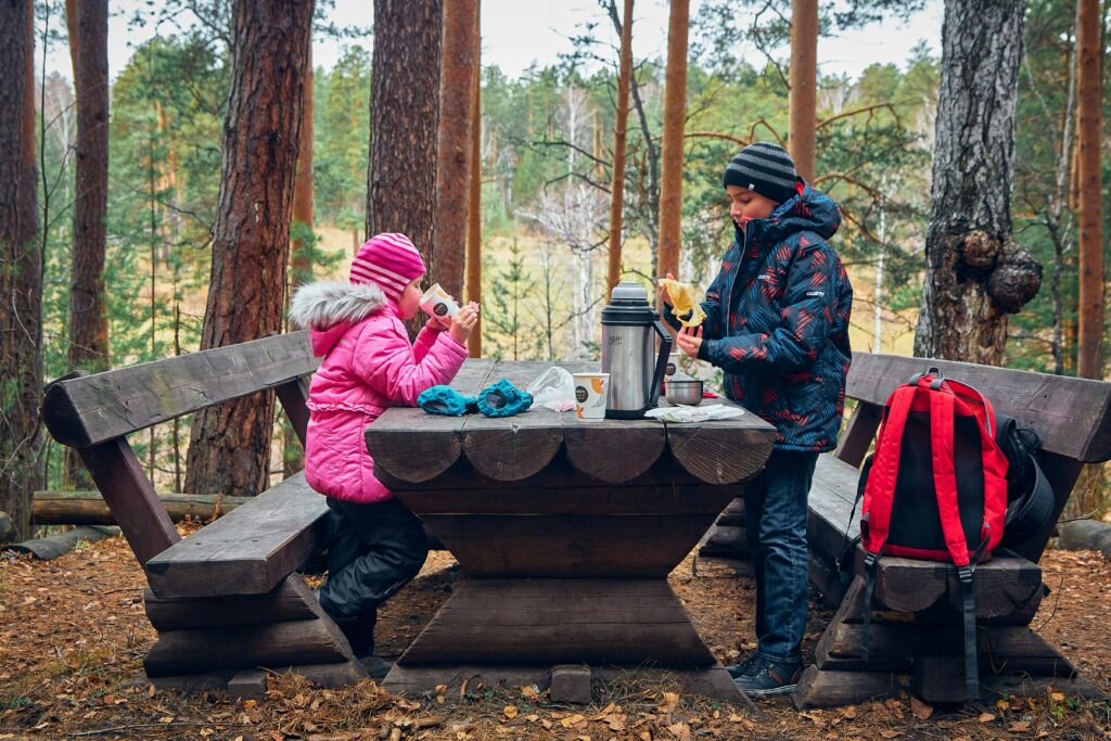 Two children enjoy a picnic in a forest, dressed in winter clothing, highlighting outdoor fun.