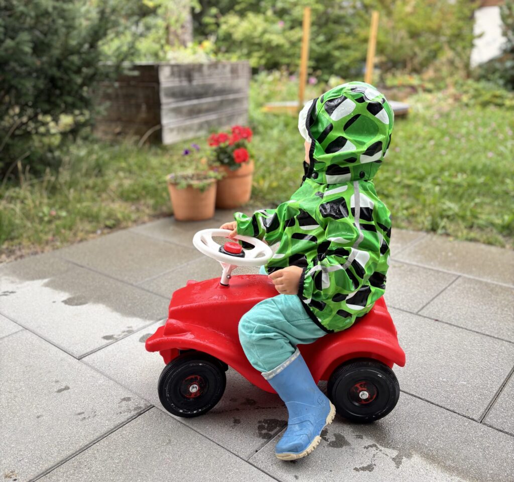 Toddler on a red Bobby Car wearing a green rain jacket and rainboots